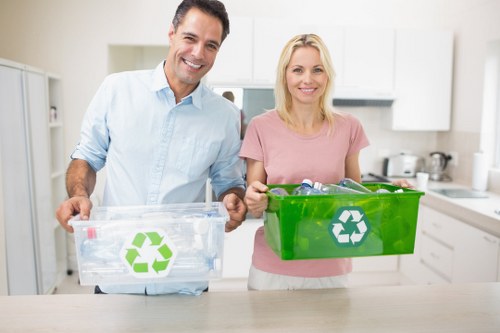 A middle-aged man with dark hair and a light blue shirt, and a woman with blonde hair wearing a pink top, standing in a modern kitchen with white cabinets and appliances in the background. They are each holding a transparent recycling bin with a green recycling symbol on the front, containing glass bottles and plastic containers, suggesting they are preparing for waste separation and recycling. The scene is well-lit with natural light, emphasizing the clean and orderly environment. This image relates to waste management and recycling services, exemplifying responsible rubbish disposal and the importance of recycling practices, which are relevant to local rubbish removal and waste clearance in areas such as Kingston upon Thames and surrounding localities.