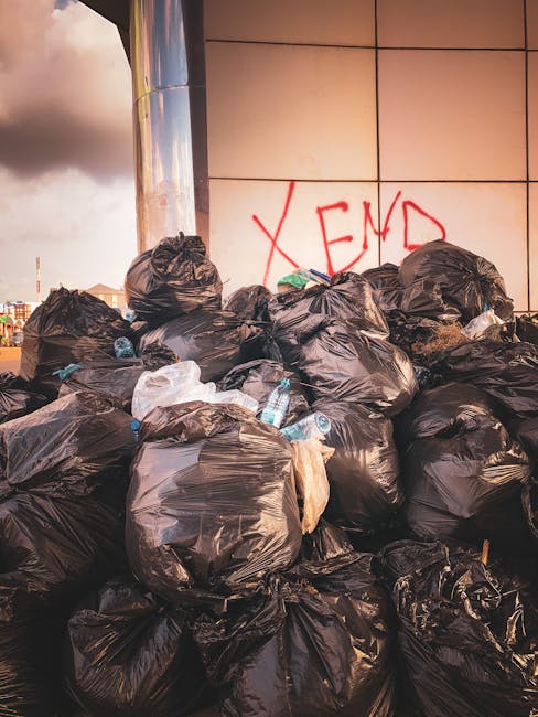 A row of tightly packed black rubbish bags piled against the exterior wall of a building, which is clad in blue-green wooden shingles. The bags are positioned close to the bottom of a small staircase 